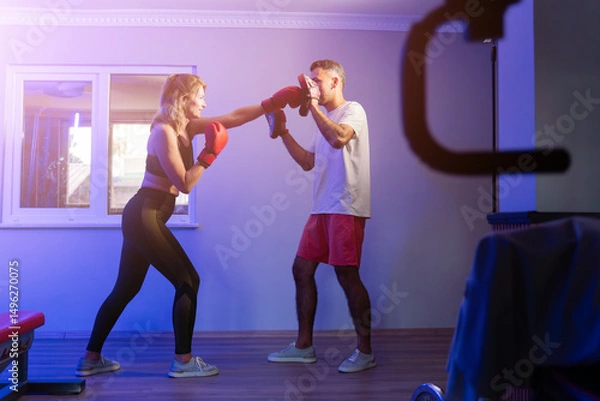 Fototapeta Female boxer training with personal coach using punch mitts in blue-lit gym