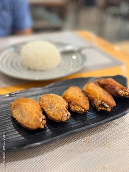 Fototapeta Golden-brown crispy fried chicken wings served on a sleek black plate, a popular appetizer or snack, with a blurred background of white rice, hinting at a full meal