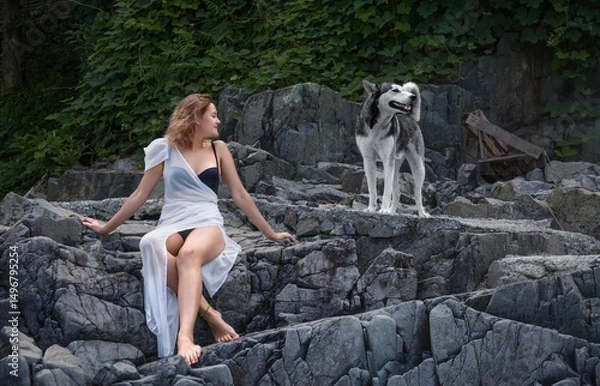 Fototapeta A young girl in a white transparent dress sits on the stone steps of a cliff and looks at a dog standing next to her