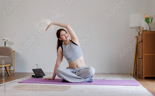 Obraz Smiling healthy Asian woman doing tutorial lesson yoga shoulder stretching at home in living room
