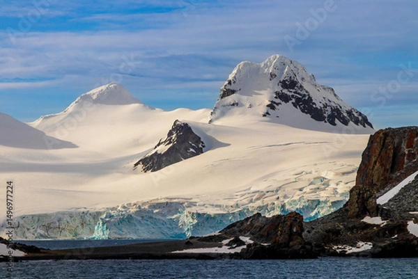 Obraz Snow covered mountain peak, early morning, Antarctic Peninsula. Rocks exposed on snowy slope. Blue ice visible. Blue sky, clouds in background. 

