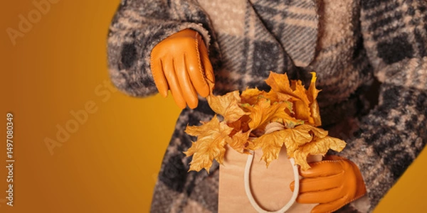 Fototapeta Woman in coat and yellow gloves holding kraft paper shopping bag full of autumn dried leaves. Autumn background