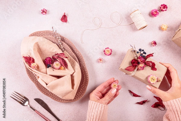 Fototapeta Process of decorating autumn table setting with dried flowers. Oval plates, cutlery, glass, gift box and natural decorations. Woman serving festive table for fall holidays, Thanksgiving Day. Top view