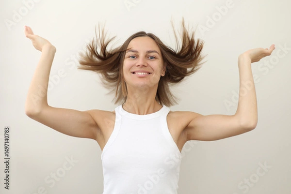 Obraz Happy young woman with flying hair smiling at camera on white backdrop. Casual natural look, raised arms, carefree emotion. Concept of freedom, fun, energy, youth branding, wellness, personal care