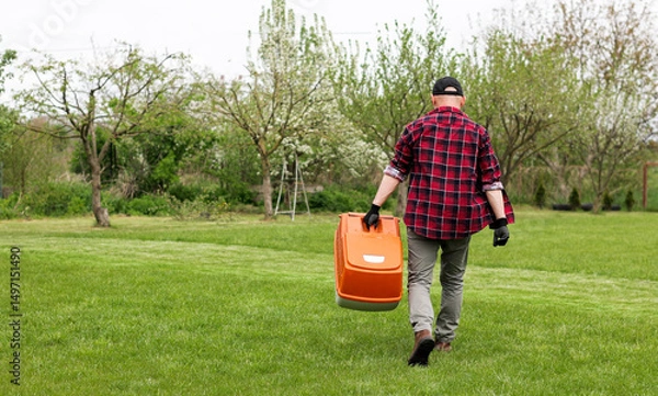 Obraz man in casual clothes holds bag with cut grass of lawn mower after mowing lawn with at own backyard. Husband takes care of garden. Modern gasoline garden equipment. Landscaping work at home