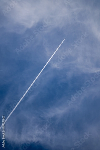 Fototapeta An airplane contrail streaks across a blue sky with wispy clouds.