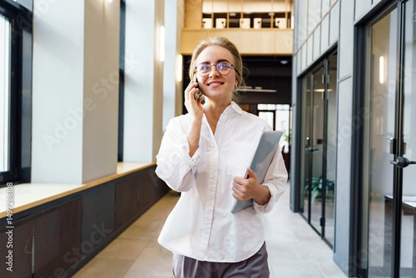 Obraz Smiling professional woman in modern office corridor engaging in a phone conversation while carrying a laptop