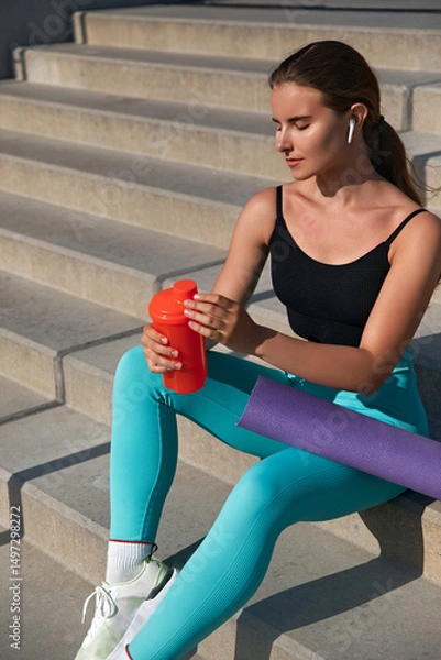 Fototapeta A young woman with a fit body sits on outdoor stairs, holding a red shaker and resting after her yoga practice. Girl enjoys the warm sunlight, embodying a lifestyle of health and fitness.
