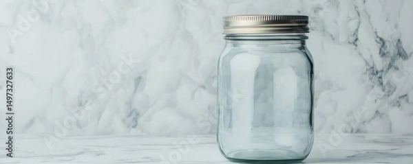 Obraz A clear, empty glass jar with a metal lid sits on a marble surface against a light background.