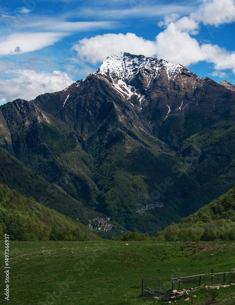 Fototapeta Charming vertical composition of Monte Legnone towering above a lush green pasture, its snow-dusted peak set against a bright blue sky with fluffy clouds. Orobie Alps, Lombardy.