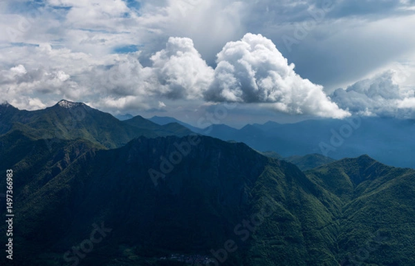 Fototapeta Atmospheric panorama of Pizzi di Parlasco, highlighting the beautiful interplay of light and shadow across verdant forested slopes, with dramatic clouds enveloping the peaks. Valsassina, Italy