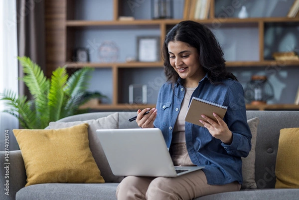 Obraz Woman attends an online meeting, smiling while holding a notepad and pen on a sofa.