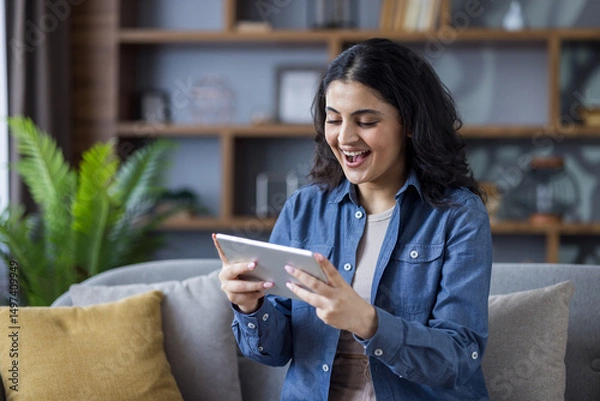 Obraz A smiling woman enjoys time on her tablet, relaxing on a comfortable sofa at home with a plant.