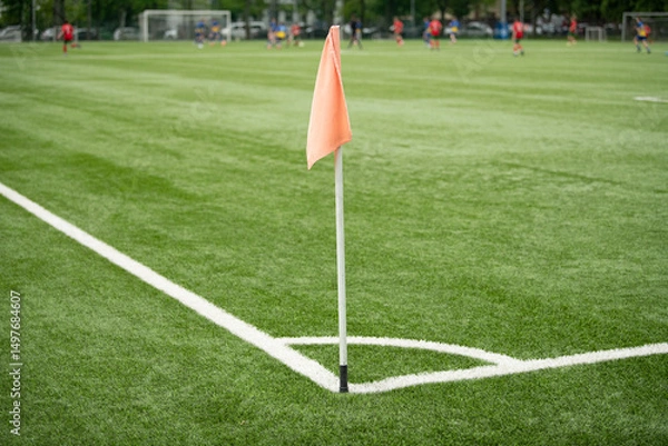 Fototapeta A vibrant corner flag stands at the edge of a manicured green soccer field. Players from two teams engage in a competitive match in the background, showcasing skillful play and teamwork