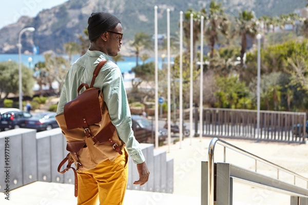 Obraz Rear view of stylish Afro American man with knapsack standing in urban setting with mountains and azure sea in background, waiting for girlfriend to have a walk on sunny day. People and lifestyle