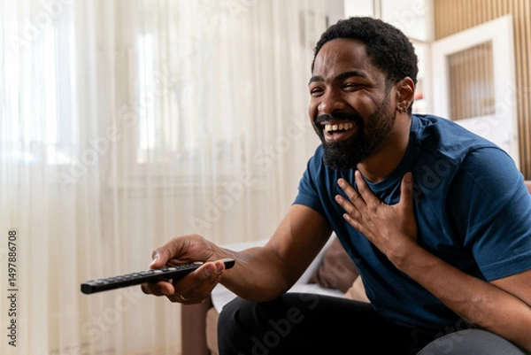 Fototapeta Young smiling happy Black man he wear casual clothes sits on sofa couch stay at home switch channel watch tv hotel flat rest relax spend free time in living room indoor. Lounge concept.