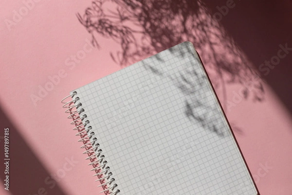 Obraz Spiral checkered white paper notepad on the pink table with leaves shadows. Copy space