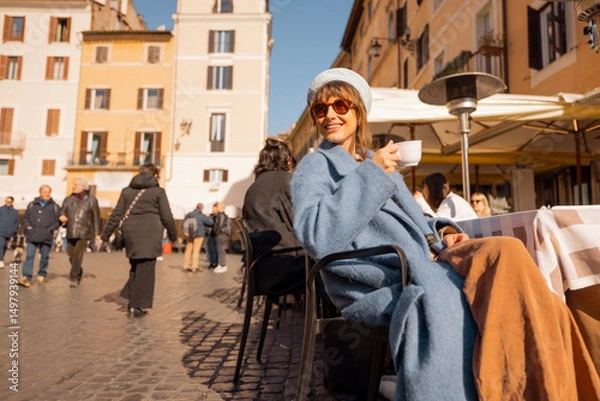 Fototapeta Smiling woman in a blue coat enjoys her coffee in a sunlit Roman square, surrounded by warm-toned buildings and people strolling by