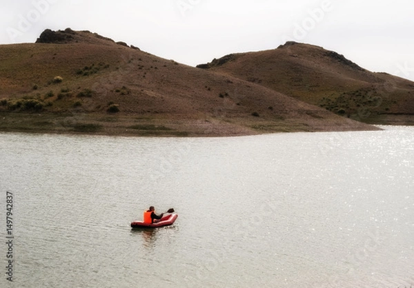 Obraz Male tourist in life jacket on red inflatable boat kayak or packraft with paddle floating on water of steppe lake with hills on sunny autumn day