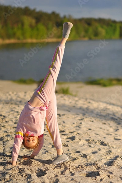 Obraz Smiling little girl playing and doing splits outdoor on sand near lake. Fitness child girl doing yoga exercise. Happy childhood
