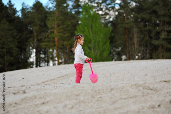 Obraz Cute child girl walking with shovel in her hand on the sand on nature background in the spring. Happy childhood, space for text