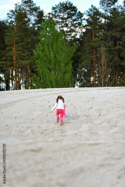 Obraz Kid girl with two ponytails running with shovel in her hand on the sand on nature background in the spring. Happy childhood, space for text