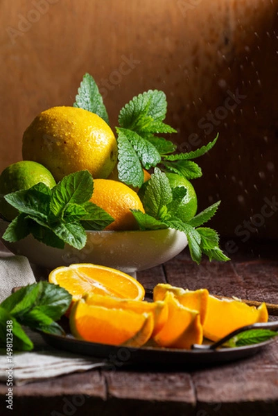Fototapeta Oranges, lemons, limes, mint and lemon balm and raindrops illuminated by the sun on a wooden background.