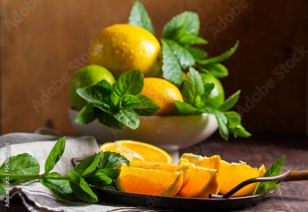 Fototapeta Oranges, lemons, limes, mint and lemon balm and raindrops illuminated by the sun on a wooden background.