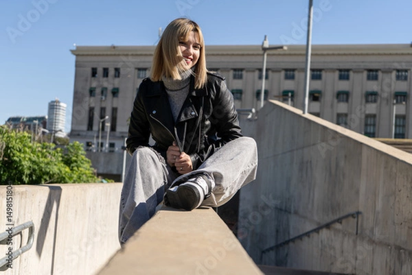 Fototapeta Stylish young woman sitting on a wall, enjoying a sunny day in the city
