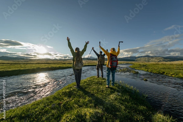 Fototapeta A group of people in bright clothes and with backpacks on their backs, stand with their hands raised in a green meadow during the golden hour. The green meadow is surrounded by rivers.