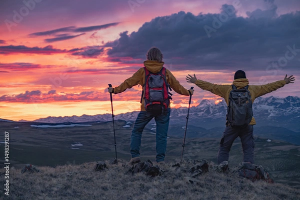Fototapeta Two travelers in bright clothes with backpacks on their backs stand on the top of a mountain during a fiery sunset, overlooking the mountains that are partially covered with snow. One of them stands w