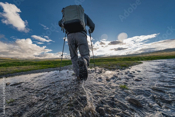 Fototapeta A photo of a man with a butt. A traveler walks on the water in trekking boots and with a backpack on his back. There is a green meadow and blue sky on the background. Waterproof boots.