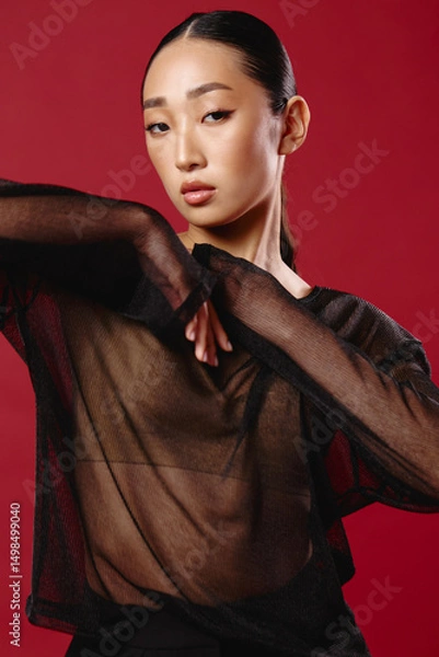 Fototapeta Fashion asian woman in sheer top and black pants poses against a red background with hands on hips and head tilted to the side
