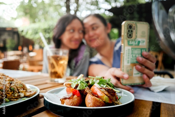 Fototapeta Young Asian mother and her teenage daughter smiling and taking selfie together using smartphone while having lunch at an outdoor restaurant. Close family relationship concept.