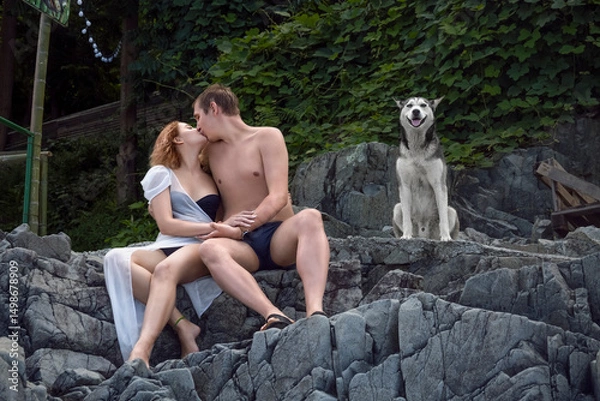 Fototapeta A young couple is sitting on the stone steps of a cliff overgrown with vines, and kissing, with a faithful dog sitting next to them