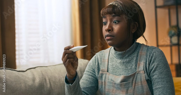 Fototapeta A woman of multi-racial background sits tiredly on her bed, checking her temperature with a thermometer. After a restless night with fever and a runny nose, she follows the doctor's instructions.