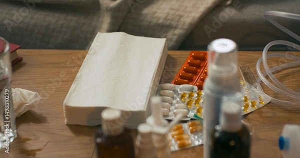 Fototapeta On the table lie paper handkerchiefs used for a runny nose, alongside various types of cold remedies including tablets, syrups, and nasal sprays arranged neatly in a sick room setting.