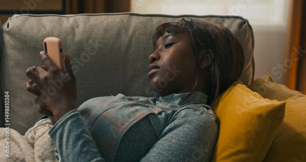Fototapeta A young woman rests at home, searching online for simple cold remedies. She scrolls through social media, mixing self-care with distraction from her symptoms and fatigue.