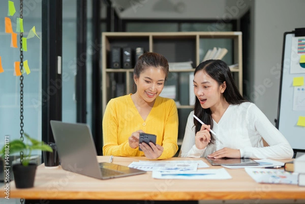 Fototapeta Two excited Asian businesswomen raising their arms and delighted at a successful company event in an office environment.