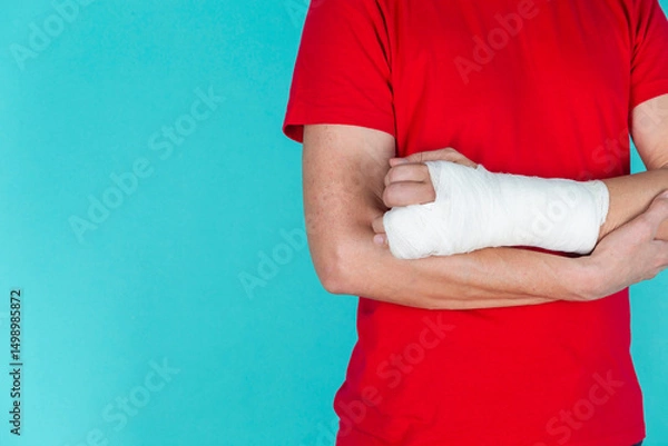 Fototapeta Medical Concepts. Closeup of Body Parts of Caucasian Man in Red Shirt with Broken Hand In Plaster and Bandaid Isolated on Turquoise Background.