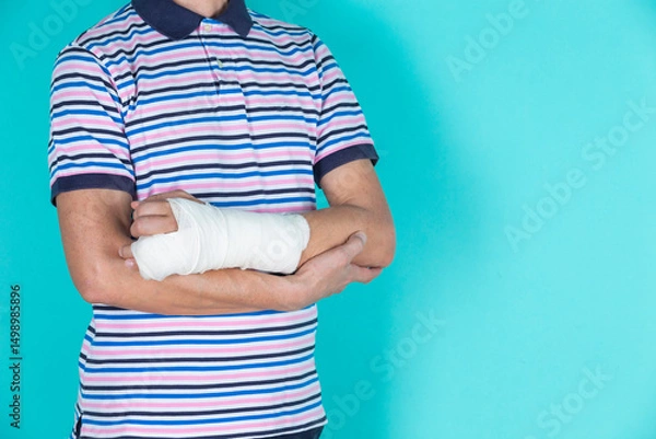 Fototapeta Medical Concepts. Closeup of Body Parts of Caucasian Man in Striped Shirt with Broken Hand In Plaster and Bandaid Isolated on Turquoise
