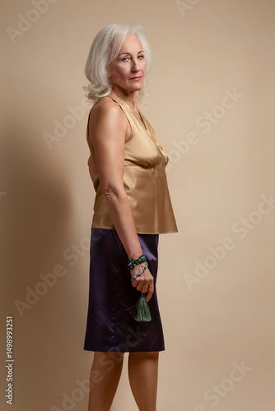 Fototapeta Ageing Concepts. Portrait of Senior Caucasian Blond Woman Posing in Casual Brown Blouse And Vilet Skirt In Studio Over Beige Background With Calm Facial Expression.
