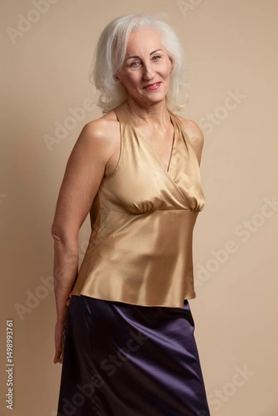 Fototapeta Portrait of Smiling Senior Caucasian Blond Woman Posing in Casual Brown Blouse And Vilet Skirt In Studio Over Beige Background With Behind Crossed Hands
