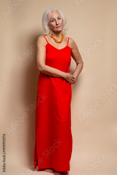 Fototapeta Ageing Concepts. One Winsome Senior Caucasian Blond Woman Posing in Red Dress In Studio Over Beige Background With Positive Facial Expression.