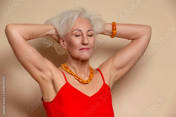 Fototapeta Ageing Concepts. Smiling Happy Winsome Senior Caucasian Blond Woman Posing in Red Dress With Lifted Hands And Eyes Closed In Studio Over Beige Background With Positive Facial Expression.