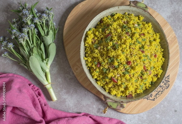 Obraz A vibrant yellow couscous salad with pomegranate seeds sits in a rustic bowl on a wooden tray, accompanied by fresh flowers. - Generative AI