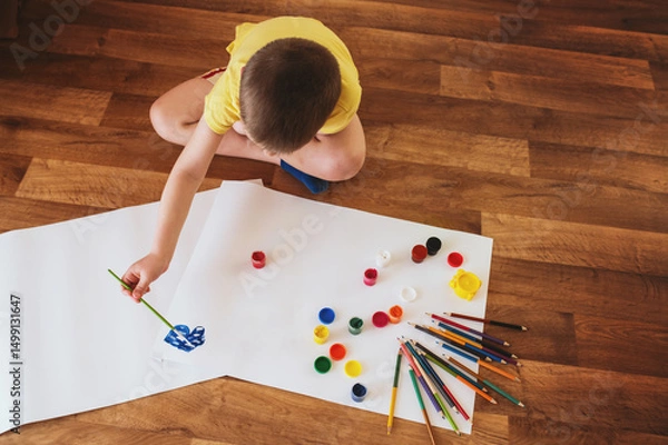 Obraz A little boy, 5 years old, a preschooler, draws with gouache paints on sheets of paper while sitting on the floor. The child is engaged in creativity, children's self-realization, independent top view