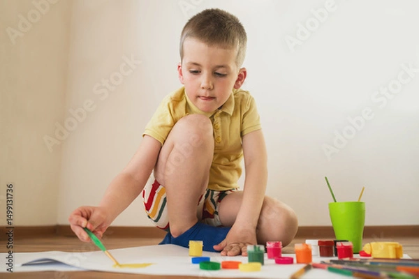 Fototapeta A little boy, 5 years old, a preschooler, draws with gouache paints on sheets of paper while sitting on the floor. The child is engaged in creativity, children's self-realization, independent, develop