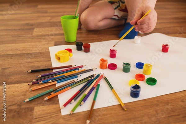 Obraz boy child preschooler, draws with gouache paints and pencil on sheets of paper while sitting on the floor. The child is engaged in creativity, children's self-realization, independent closeup no face