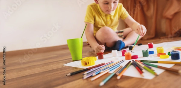 Obraz A little boy, 5 years old, a preschooler, draws with gouache paints on sheets of paper while sitting on the floor. The child is engaged in creativity, children's self-realization, copy space banner
